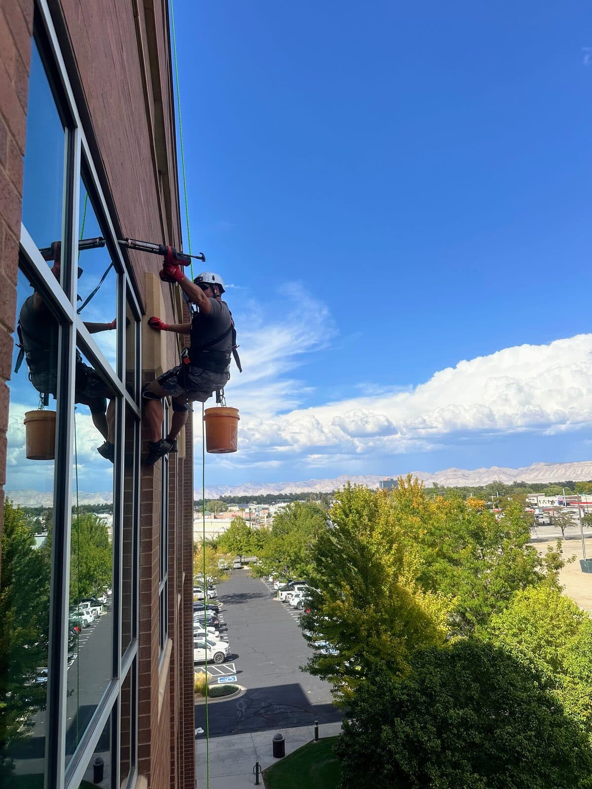 Rope-access technician on a sunny glass building with blue sky, mountains, and an orange bucket, reflection in the windows