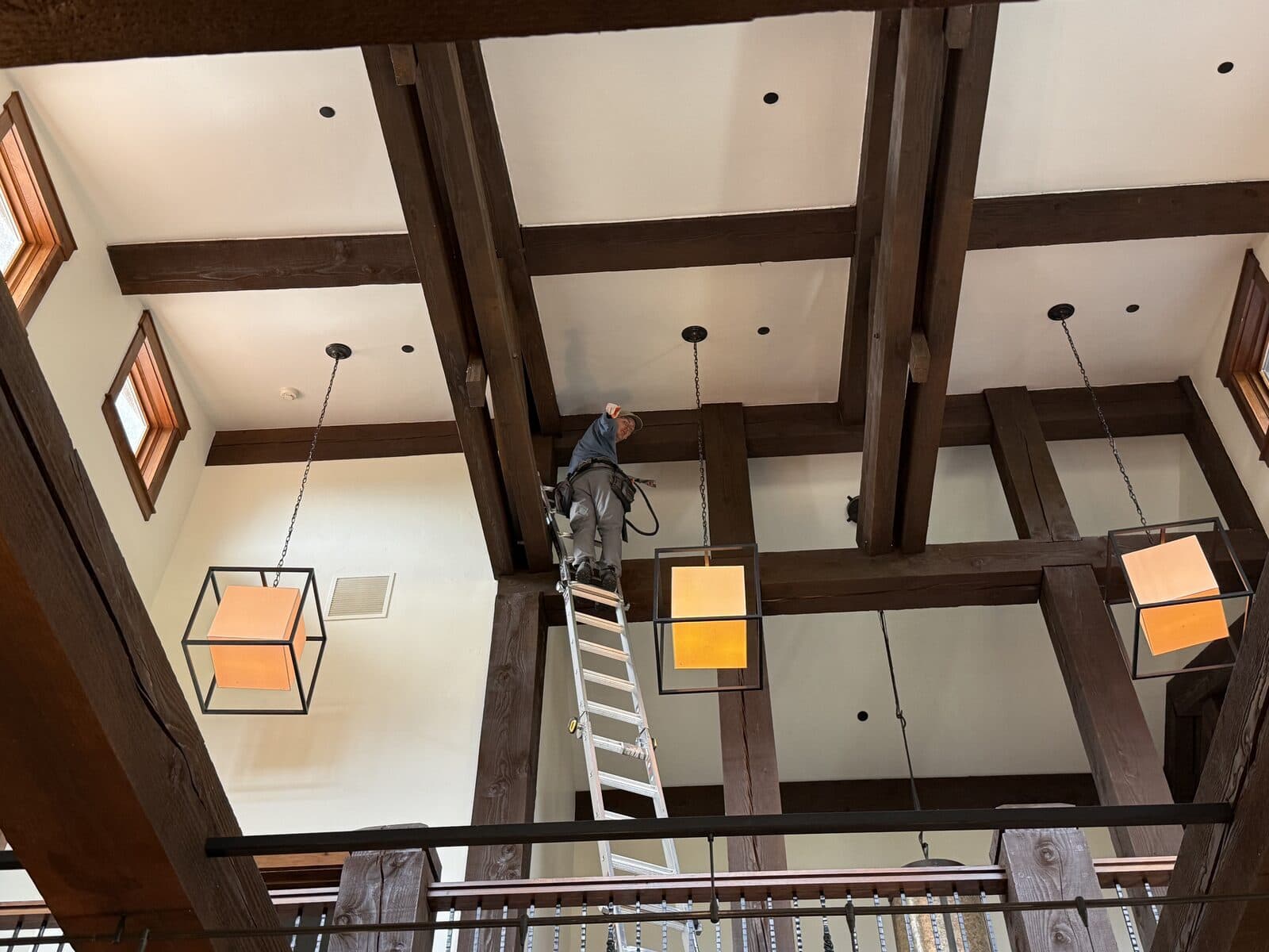 Tall interior with dark wood beams and cube lights, technician on a tall ladder at the ceiling