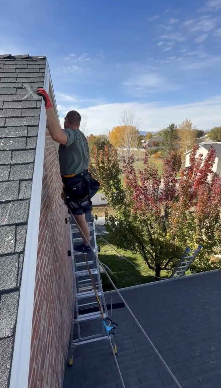 Technician on a tall ladder at a brick gable and white fascia under a blue sky
