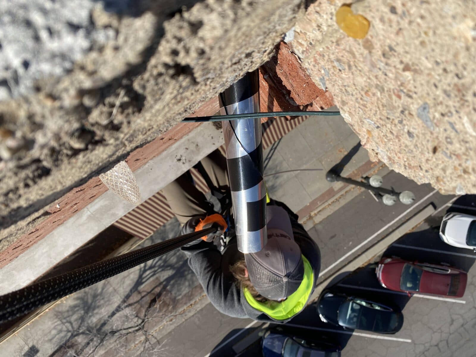 Rope-access technician installing a one-way exclusion tube on a multi-story facade above a parking lot
