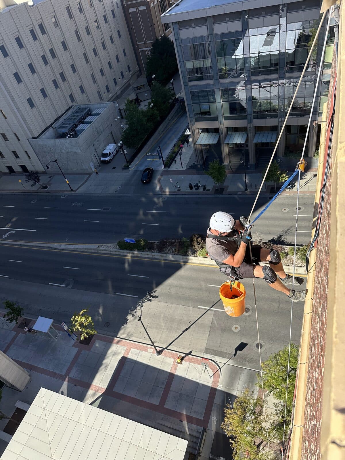 High-rise rope rappel down a brick facade over a city street, orange bucket hanging from the harness